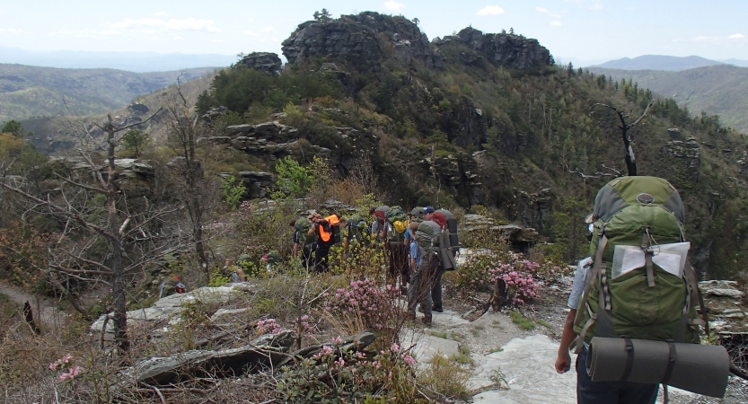 A group of backpackers hike through rocky terrain toward a large rock formation. 